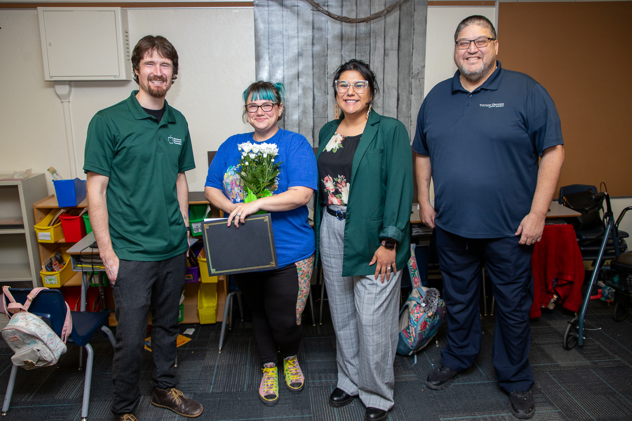 A woman in a blue shirt proudly holds her award certificate surrounded by two men and a woman