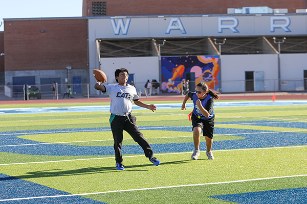 A boy in a gray shirt throws a football as a girl in a blue jersey runs towards him