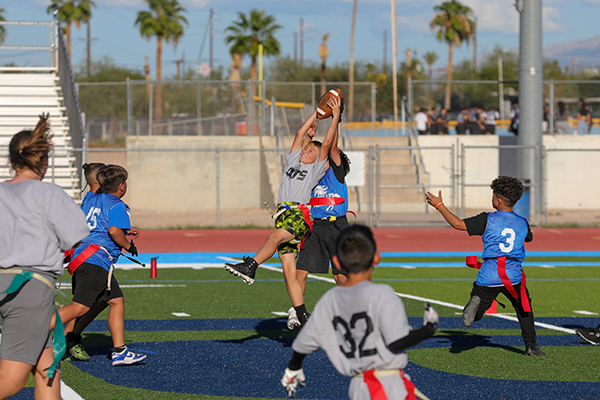 A boy in a gray shirt leaps up to grab a football in the air away from an opponent
