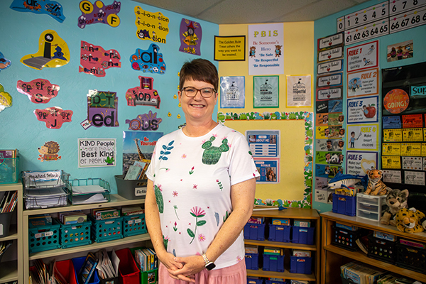 A woman with short brown hair, glasses and a cactus shirt smiles in front of colorful classroom walls